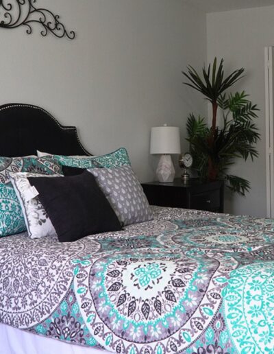 A cozy bedroom with an ornate black headboard and a bed covered in a teal and black patterned comforter. a bedside table with a lamp, a plant in the corner, and an open door are visible.
