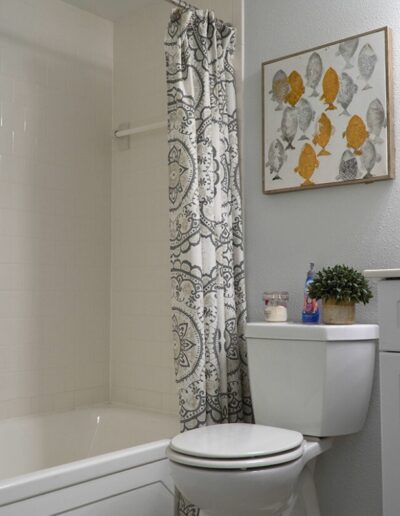 A small, neatly organized bathroom featuring a white tub with a patterned shower curtain, a toilet, and a wall-mounted art piece with floral prints. decor includes a potted plant and candles on a shelf.