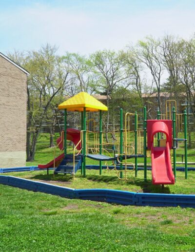 A colorful playground with a red slide, yellow canopy, and green bars, surrounded by grass and a building on the left.