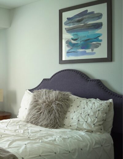 A cozy bedroom corner featuring an elegantly tufted gray headboard with a white comforter, a fluffy gray pillow, and a modern abstract blue painting hanging on the wall.