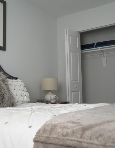 A cozy bedroom featuring a gray upholstered headboard with white bedding, a plush gray throw, a bedside lamp, and decorative wall art, with an open closet door in the background.
