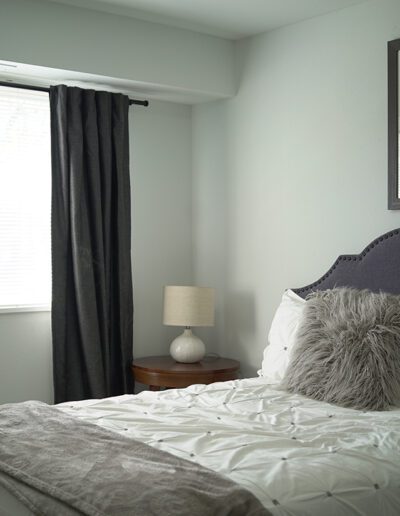 A cozy bedroom with a gray upholstered headboard, white bedding, and a fur pillow. a lamp sits on a bedside table, and abstract art hangs above the bed. light filters in through a window with blinds and curtains.