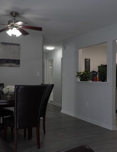 A modern dining area with a dark wood table and chairs under a ceiling fan. adjacent is an open kitchen, visible through a white framed doorway. neutral tones and a wall painting enhance the space.