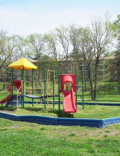 A colorful playground with a blue safety border, located at North Hills Apartments, featuring a yellow and green structure with slides and climbing areas, set against a backdrop of trees and apartment buildings.