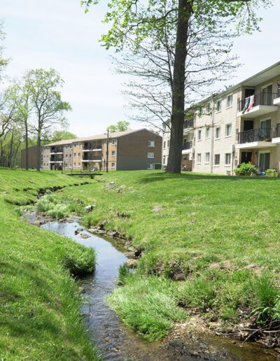 A serene North Hills apartment complex with brick buildings overlooking a small, winding creek, surrounded by lush green grass and scattered trees on a sunny day.