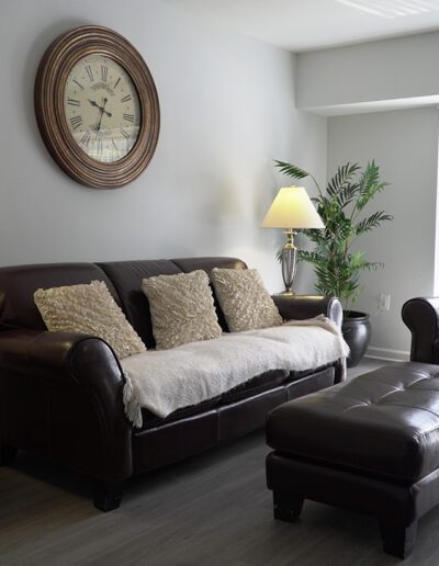 A cozy living room featuring a dark brown leather sofa with beige cushions, a matching armchair and ottoman, two lit lamps, and a large clock on a gray wall.