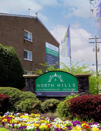 An entrance sign of North Hills Apartments For Rent with contact information, adorned by colorful flowers and green shrubs, under a sunny sky with a brick building and flying banners in the background.
